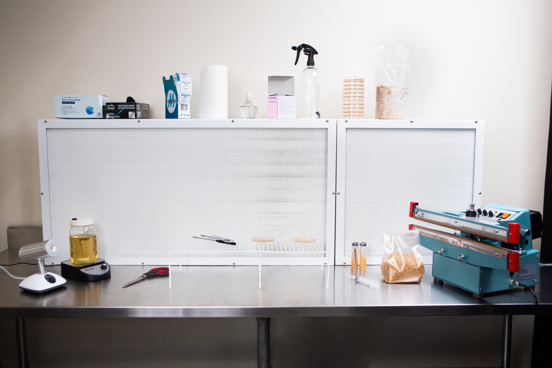 A clean lab workspace features a Laminar Flow Hood HEPA Airfilter H14 with pre-filter 60x60x35cm, equipment, petri dishes, jars, and a vacuum sealer on a stainless steel table.