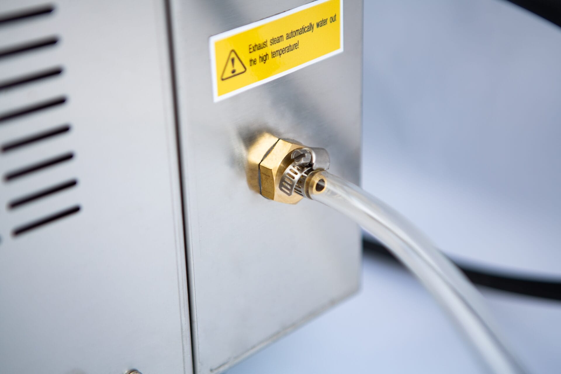 Clear tube connected to a brass fitting on a metal machine; warning label about high temperature nearby.