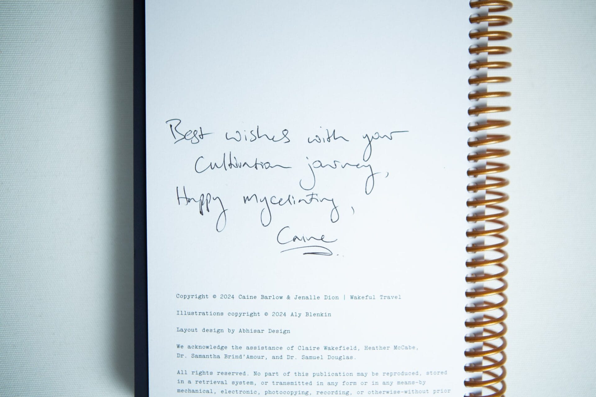 A handwritten note on a book page reads: Best wishes with your cultivation journey, happy myceliating, Carrie—inside the "Wakeful Mushroom Cultivation Journal" by Australian mycologist Caine Barlow.