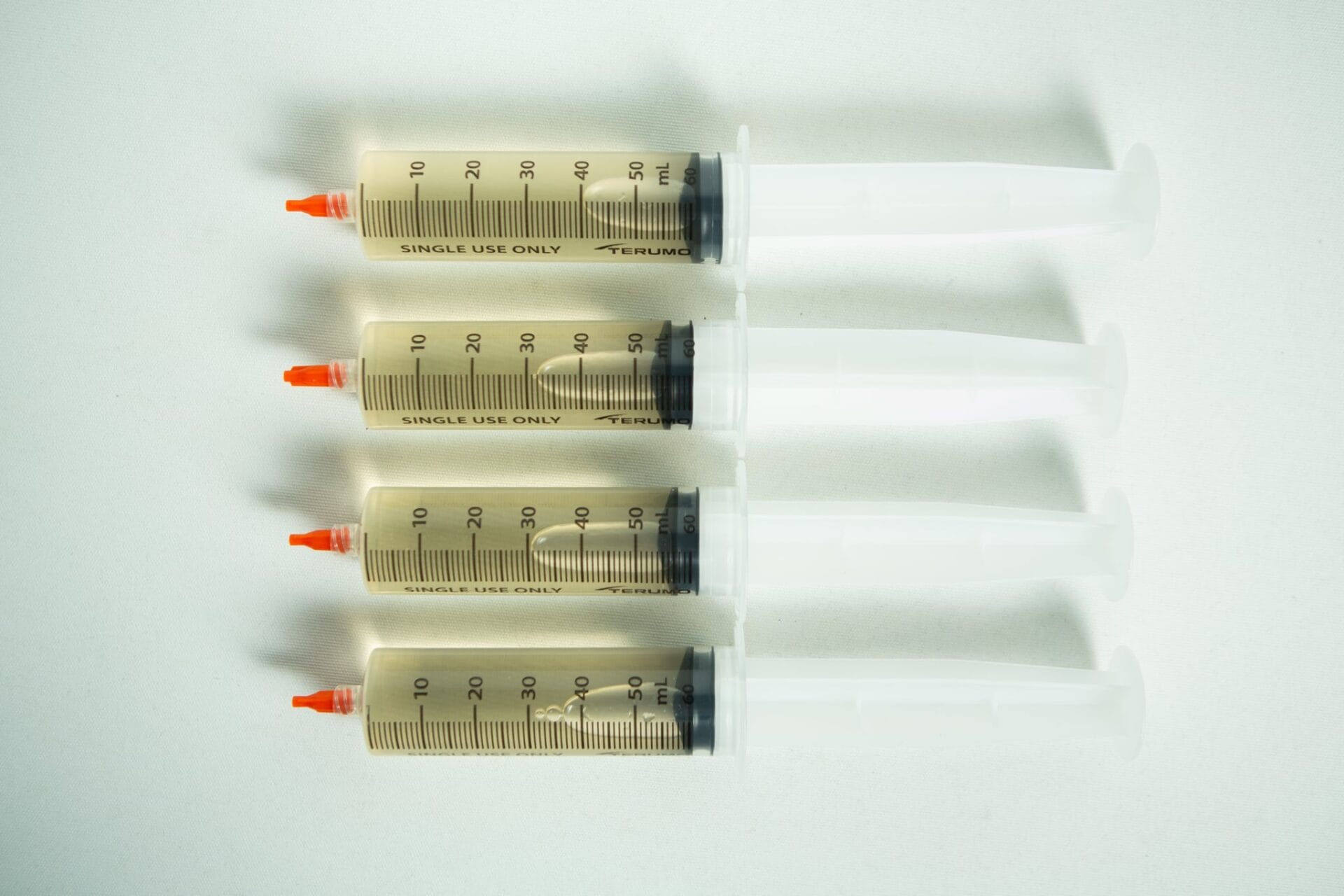 Four large syringes with orange caps from the Liquid culture to Liquid culture expansion Kit are lined up horizontally on a white surface.