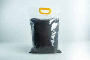 A Manure Bag with a yellow handle, made of clear plastic and filled with dark soil, displayed against a white background.