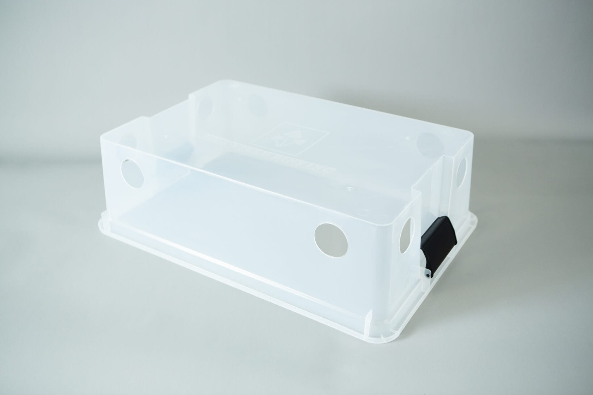 A clear plastic monotub kit with circular holes and a black catch, designed for growing mushrooms, is placed on a light grey surface.