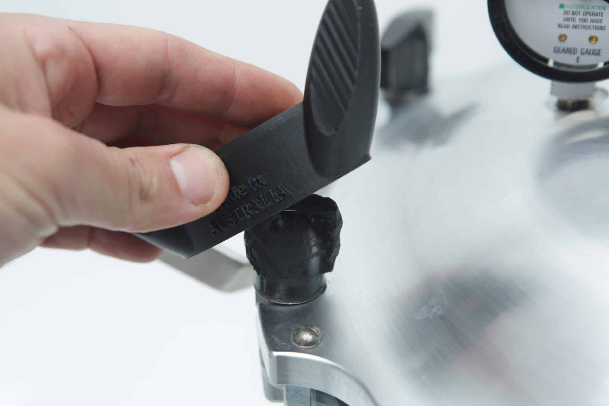 A hand removes a black safety valve from the lid of an All American Tool stainless steel pressure cooker.