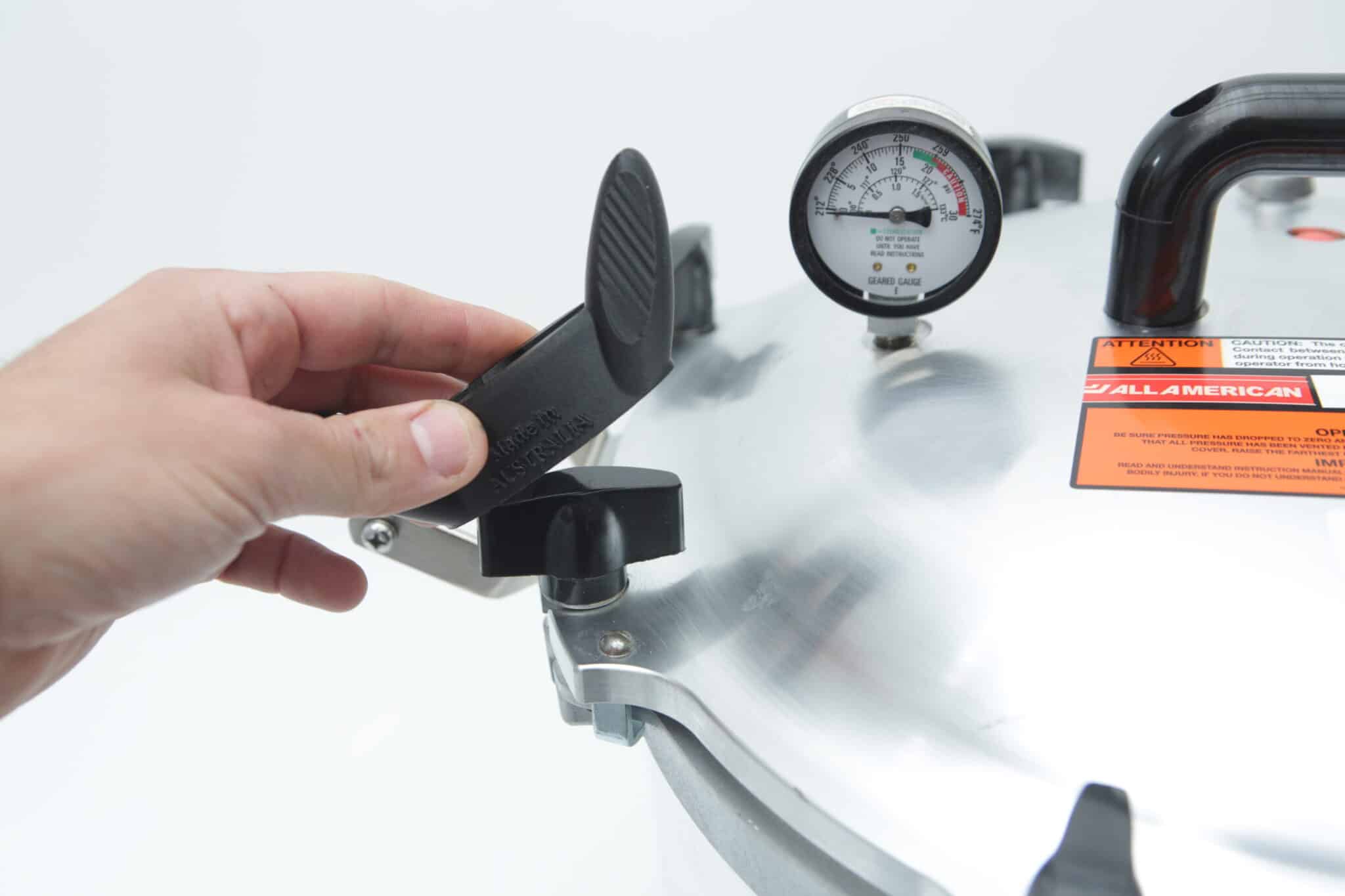 A hand turns the locking mechanism on the lid of an All American Tool pressure cooker with a pressure gauge.