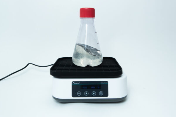 A clear bottle with liquid sits on an electronic lab shaker against a white background.