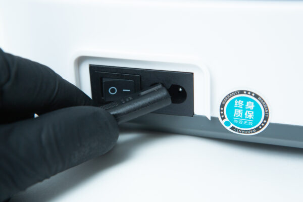 A gloved hand plugs a power lead into a device with an on/off switch and a warranty sticker.