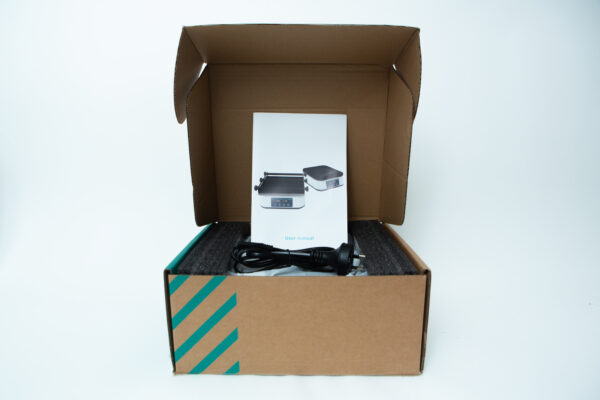 Open cardboard box containing an electronic device, user manual, and a power lead on foam padding.
