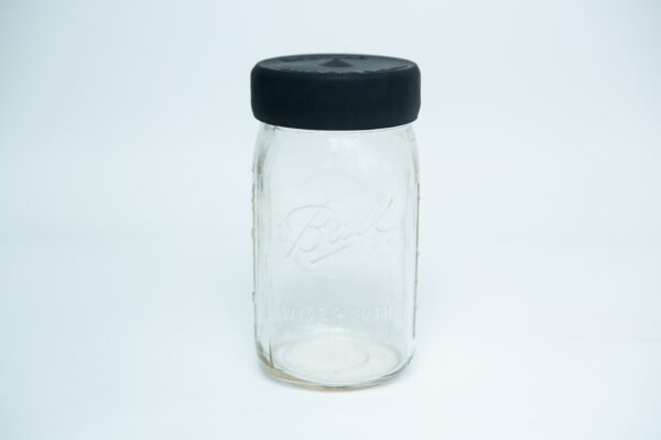 A Silicone Kilner Jar Rubber Lid for wide mouth jars is shown on a clear jar with a black lid, set against a plain white background.