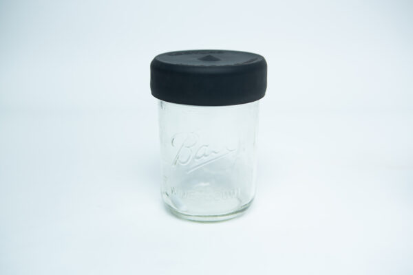 A clear glass jar with a wide mouth is displayed against a plain white background, featuring the Silicone Mason Jar Rubber Lid in black.