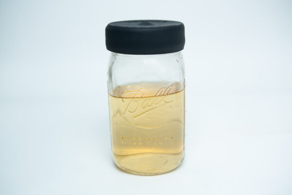 A wide-mouthed glass mason jar with a silicone mason jar rubber lid sits half-filled with light brown liquid on a white background.
