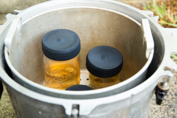 Three wide-mouth jars with silicone Mason jar lid covers (autoclavable) are placed inside a large metal pressure cooker, viewed from above.